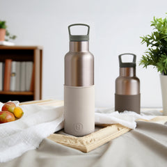Two metallic water bottles on a table with a fruit bowl and plant in the background.