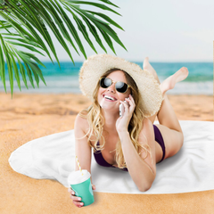 Woman in a bikini and sun hat lying on a towel at the beach, holding a drink and phone.