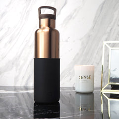 Rose gold water bottle with black sleeve on a marble surface with a candle and decorative object in the background.