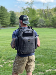Man wearing a gray backpack outdoors on a grassy field with trees in the background