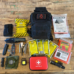 Collection of survival gear including a black backpack, first aid kit, and various tools on a wooden surface.