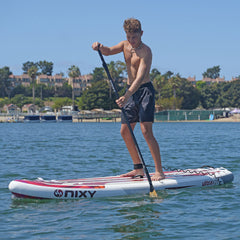 Person paddleboarding on a Nixy board in a body of water with trees and buildings in the background.