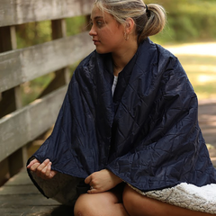 Person sitting on a wooden bench wearing a navy blue poncho by a body of water.