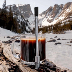 Clear mug with coffee and a metal straw on a wooden log in a snowy mountain landscape