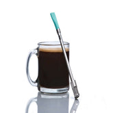 Clear glass mug with coffee and a metal straw with turquoise top on a white background