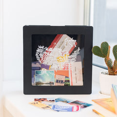 Black shadow box with travel-themed cards on a white surface with a cactus plant in the background.