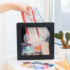 Black photo frame with a hand placing travel tickets inside, surrounded by a cactus plant and other items.