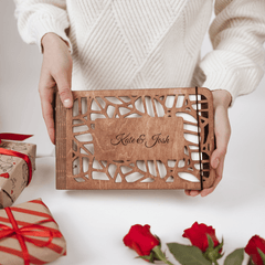 Person holding a wooden laser-cut card with 'Kate & Josh' engraving, surrounded by gift boxes and roses.
