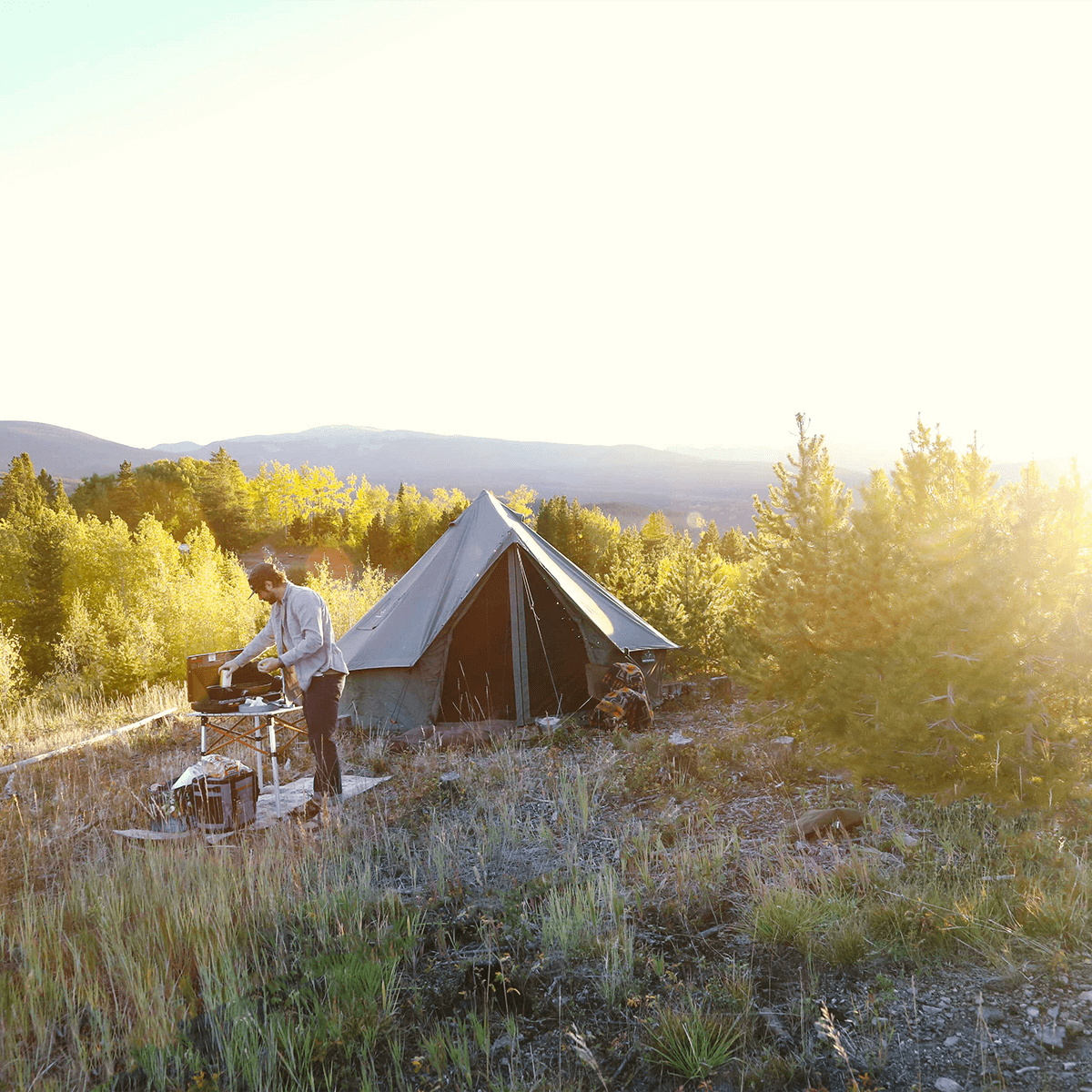Person standing next to a tent in a forested area with mountains in the background