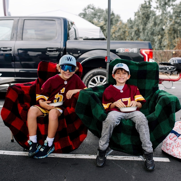 Two children sitting in colorful chairs outdoors, possibly at a tailgate event.