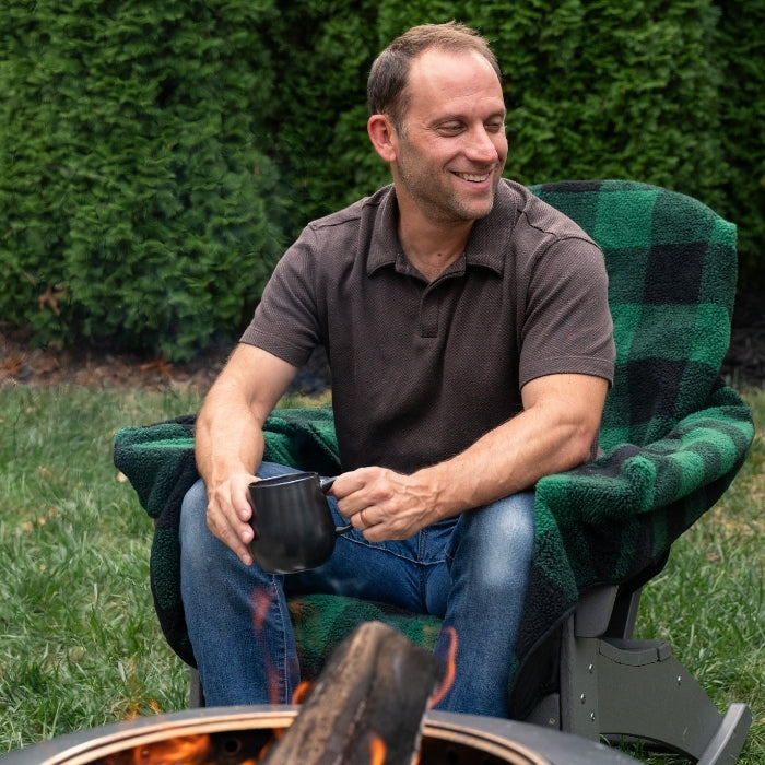 Man sitting in a green and black chair by a fire pit, holding a mug.