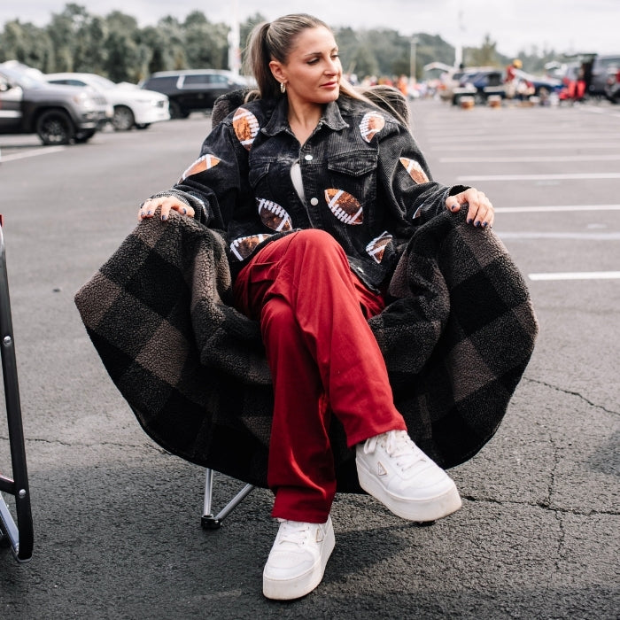 Woman sitting on a black and gray checkered chair in a parking lot wearing a patterned jacket, red pants, and white sneakers.