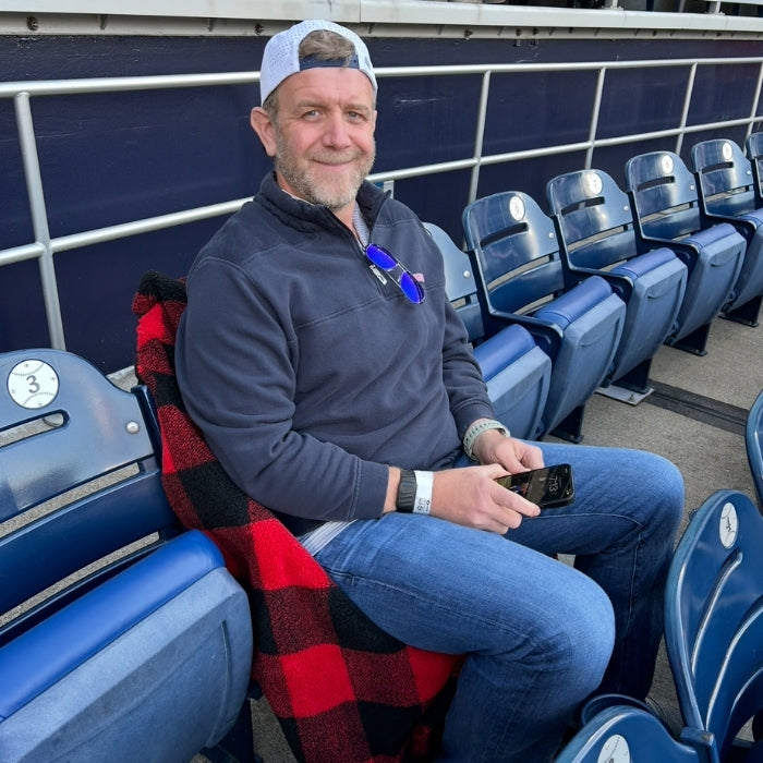 Man sitting in a stadium seat with a red and black checkered blanket, holding a phone.
