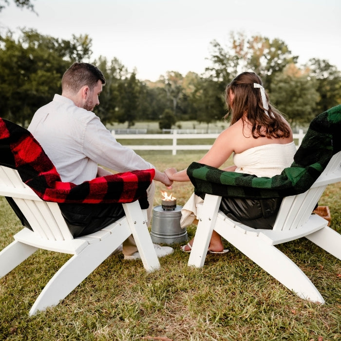 Two people sitting in Adirondack chairs by a fire pit in a park-like setting.