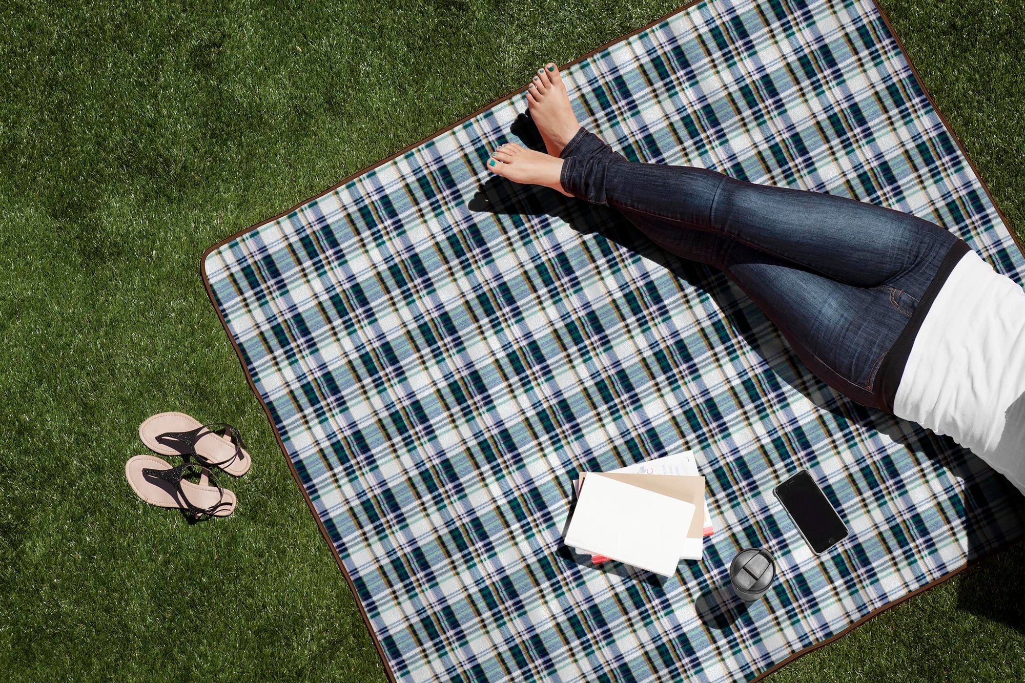 Person lying on a plaid picnic blanket on grass with a smartphone and sunglasses.