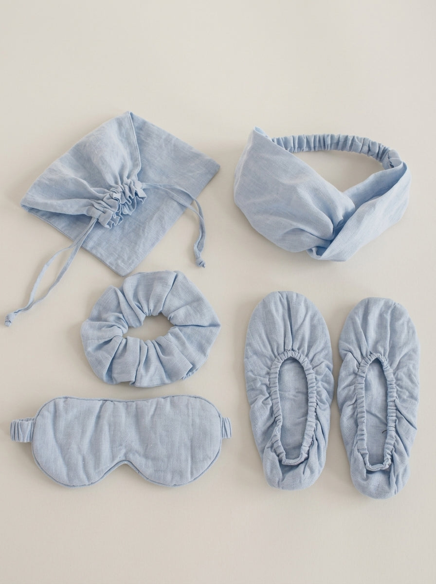 Set of light blue sleep accessories including a bonnet, headband, eye mask, and slippers on a beige background.