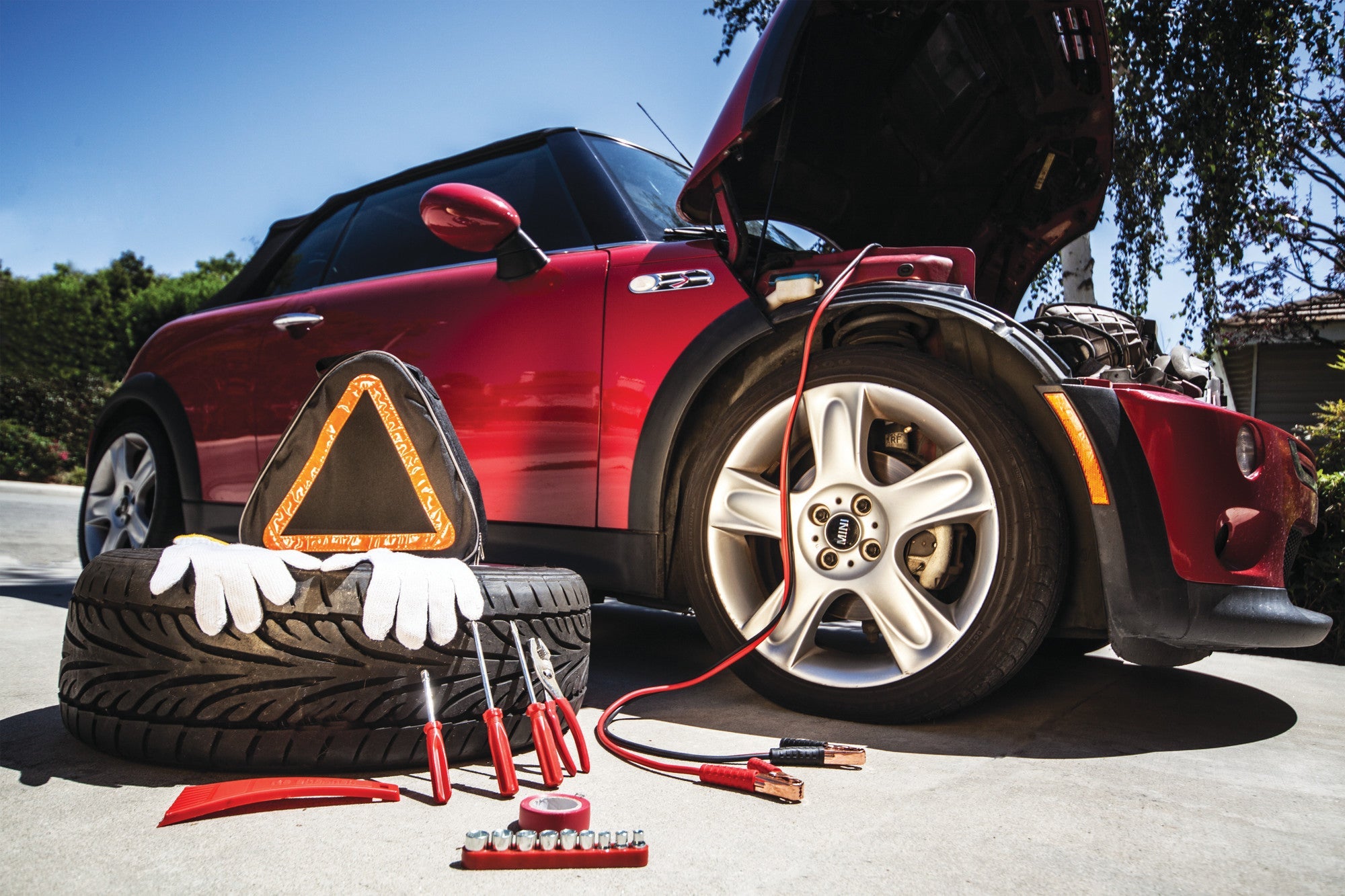 Red car with open hood, tire, tools, and warning triangle on a sunny day.