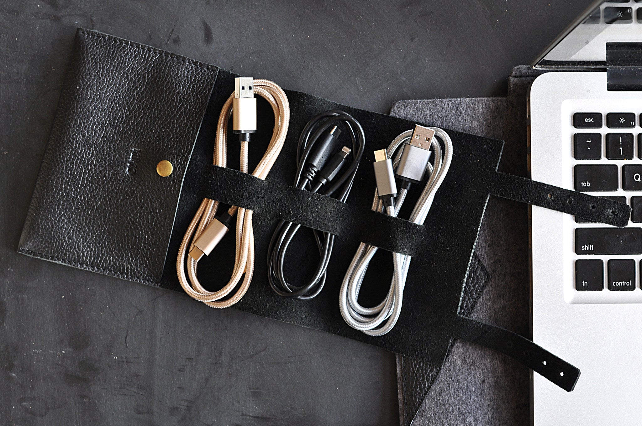 Black leather cable organizer with USB cables on a dark surface next to a laptop.