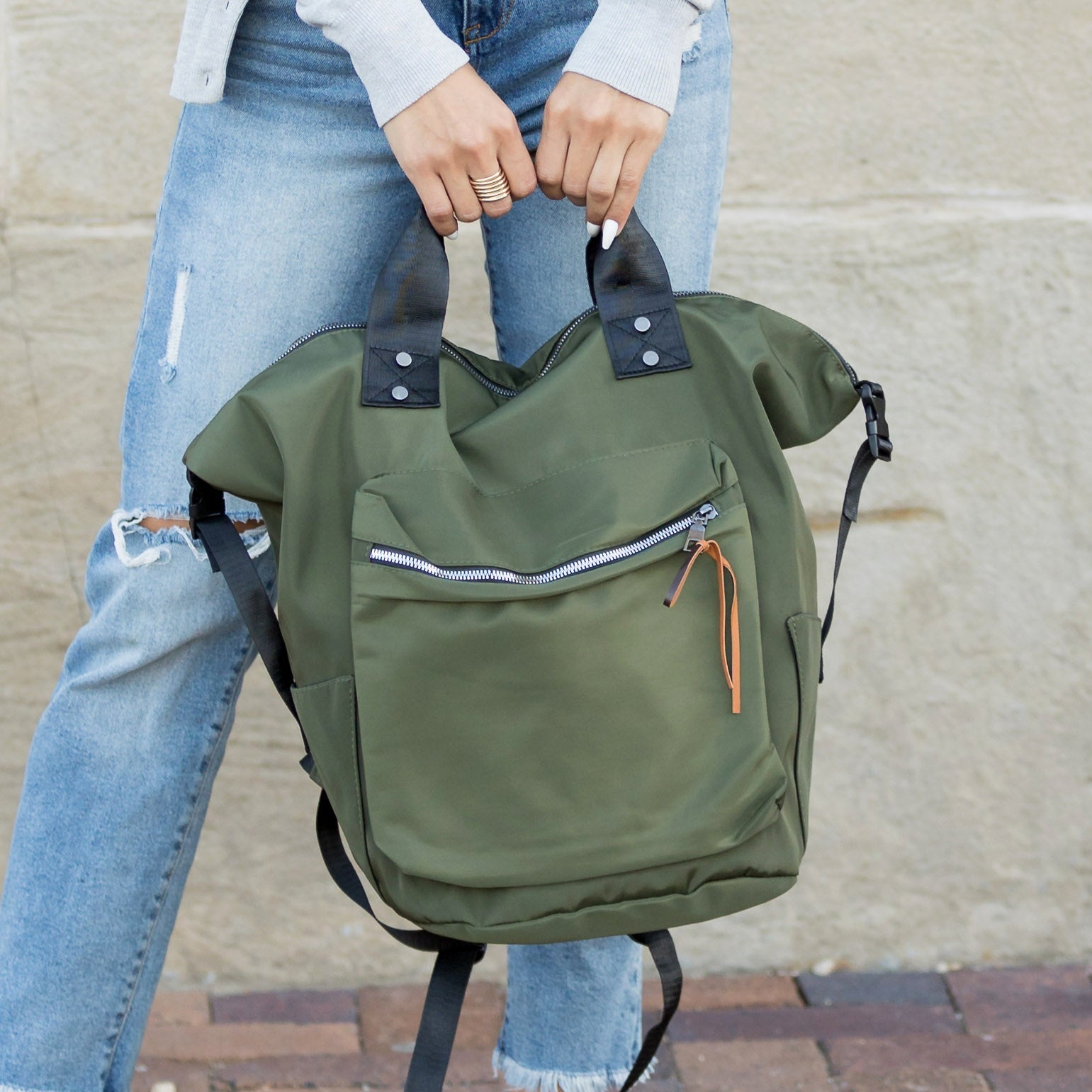 Green backpack held by a person wearing blue jeans against a beige wall.