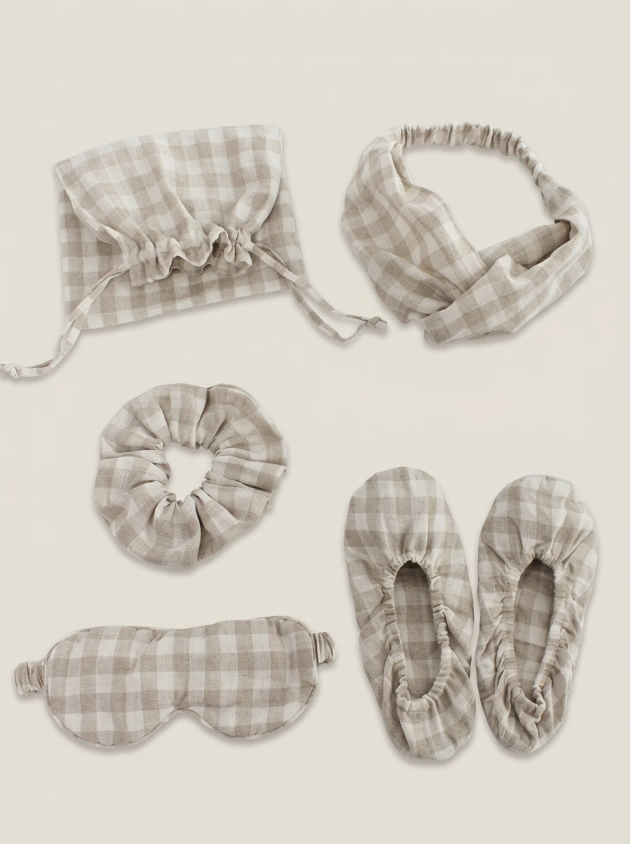 Set of gray plaid sleep accessories including a bonnet, headband, eye mask, slippers, and a drawstring bag on a beige background.