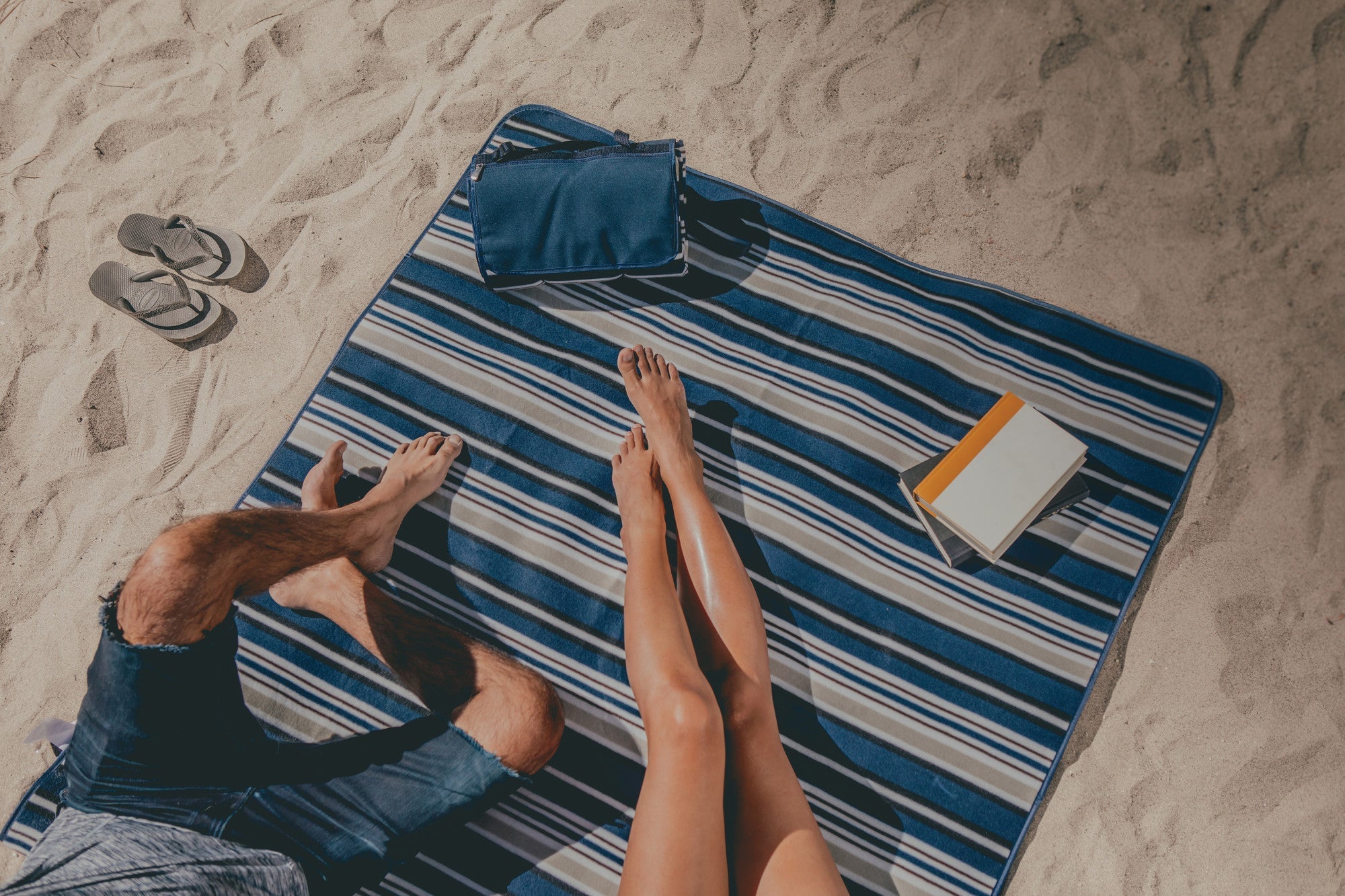 Two people lying on a striped beach towel with a book and bag on a sandy beach.