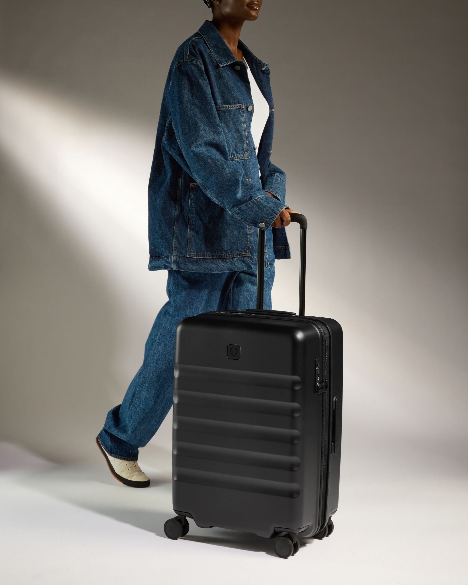 Person in blue denim outfit pulling a black suitcase against a neutral background