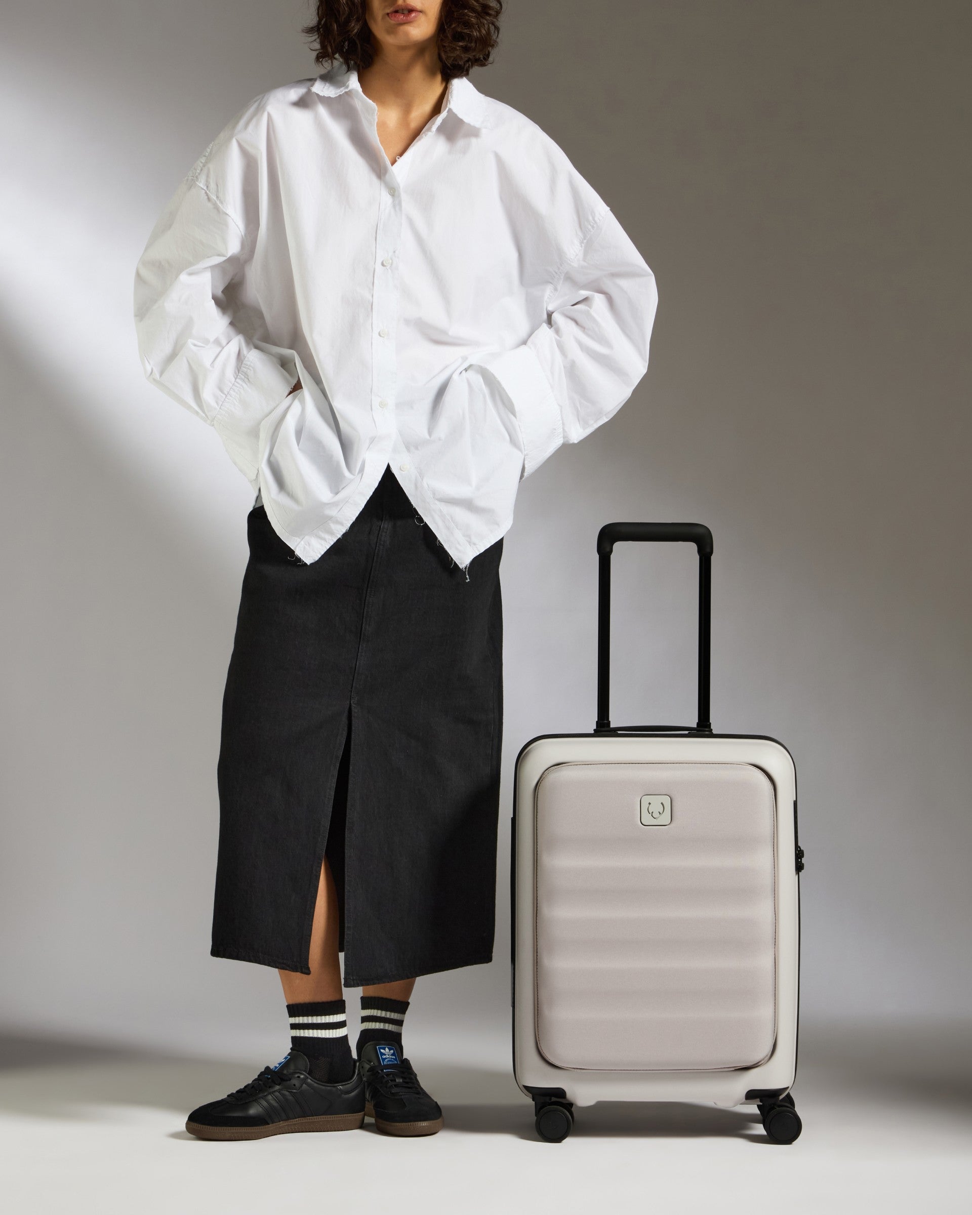 Person wearing a white shirt and black pants standing next to a silver suitcase on a gray background