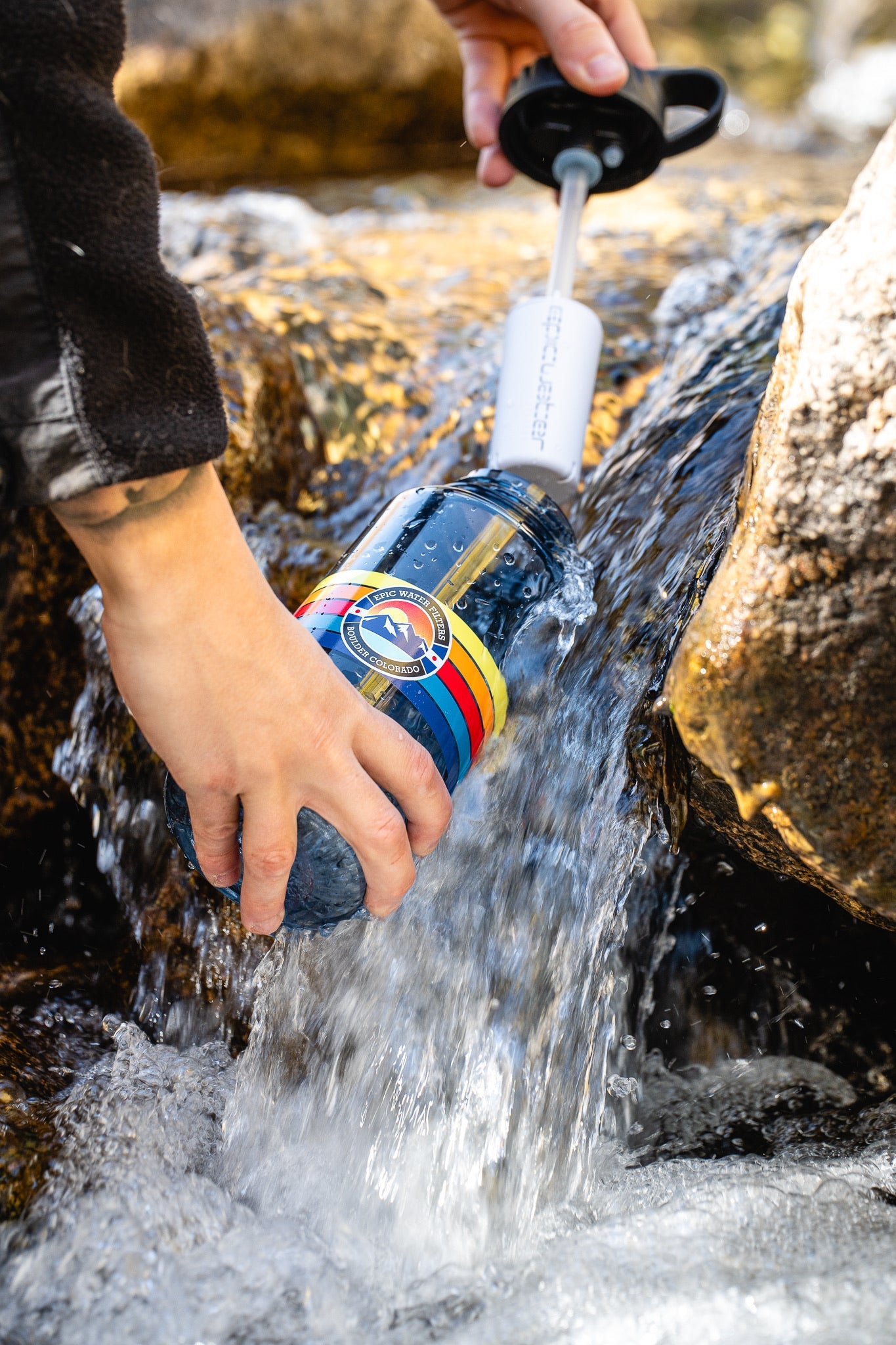 Person filling a colorful water bottle with water from a stream.