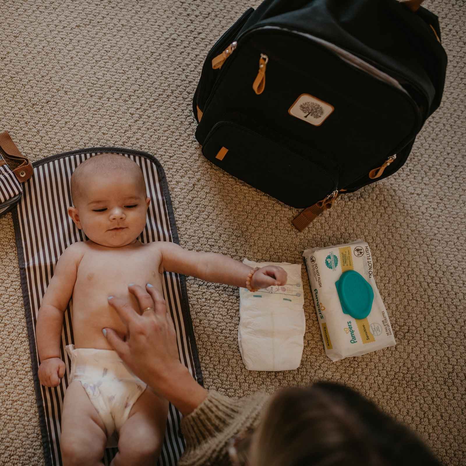 Baby lying on a changing mat with a diaper, wipes, and a backpack in the background.