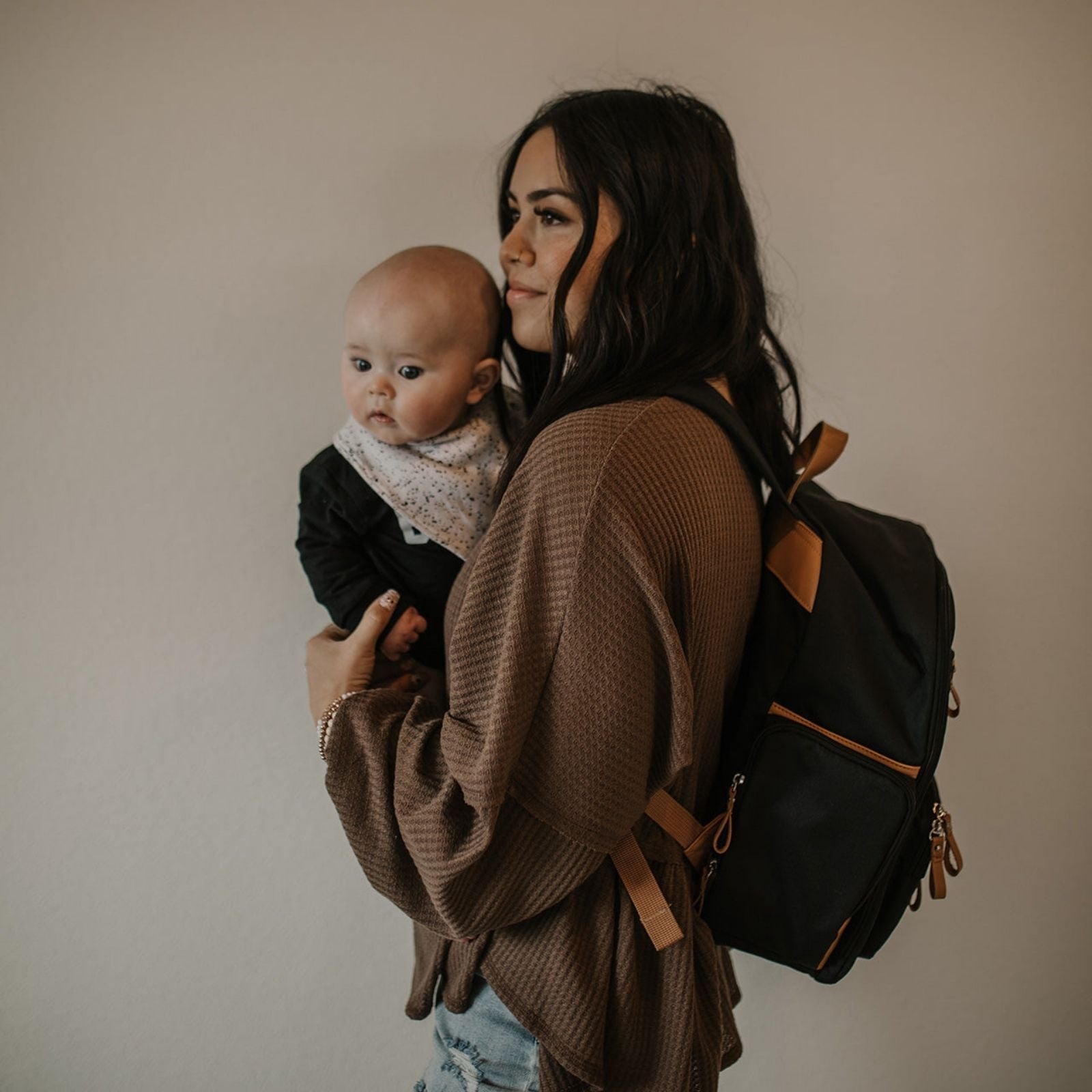 Woman holding a baby and wearing a backpack against a plain background