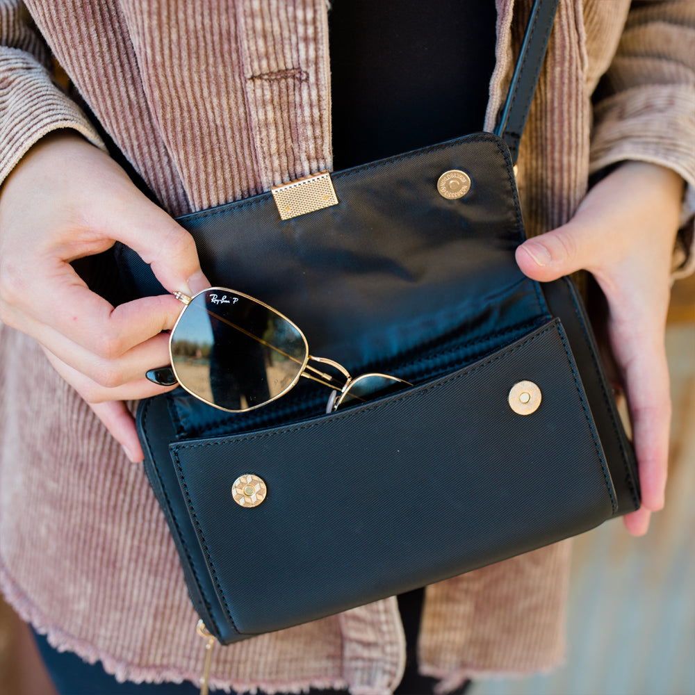 Person holding a black handbag with sunglasses inside, wearing a brown jacket.