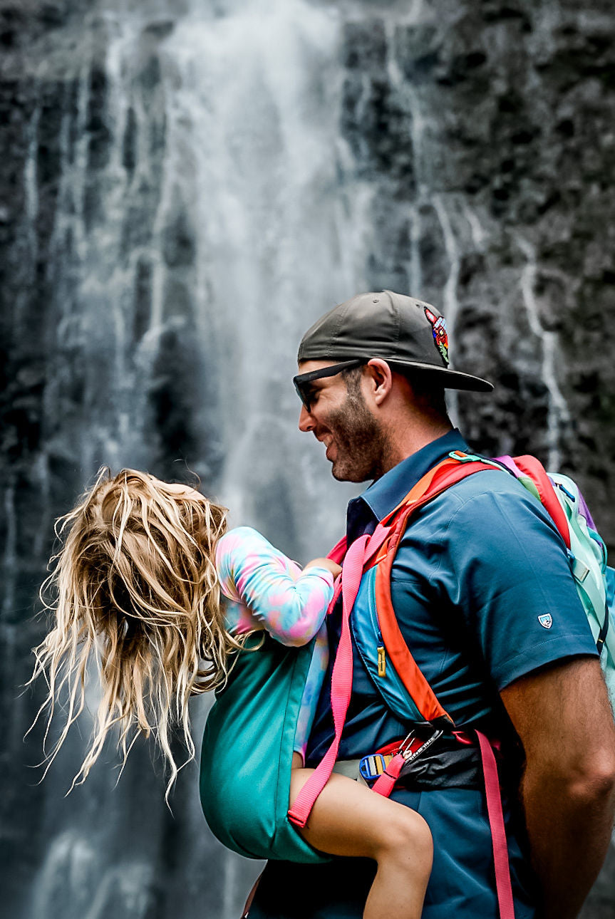 Man carrying a child in a sling with a waterfall in the background