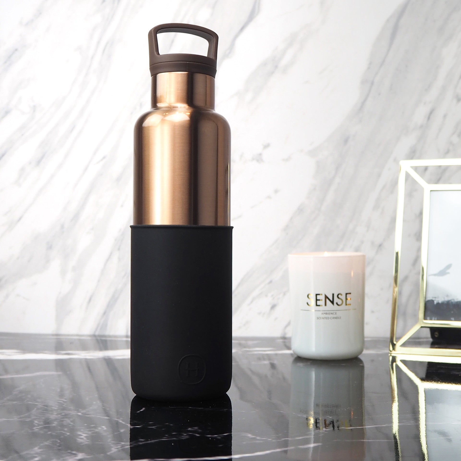 Rose gold water bottle with black sleeve on a marble surface with a candle and decorative object in the background.
