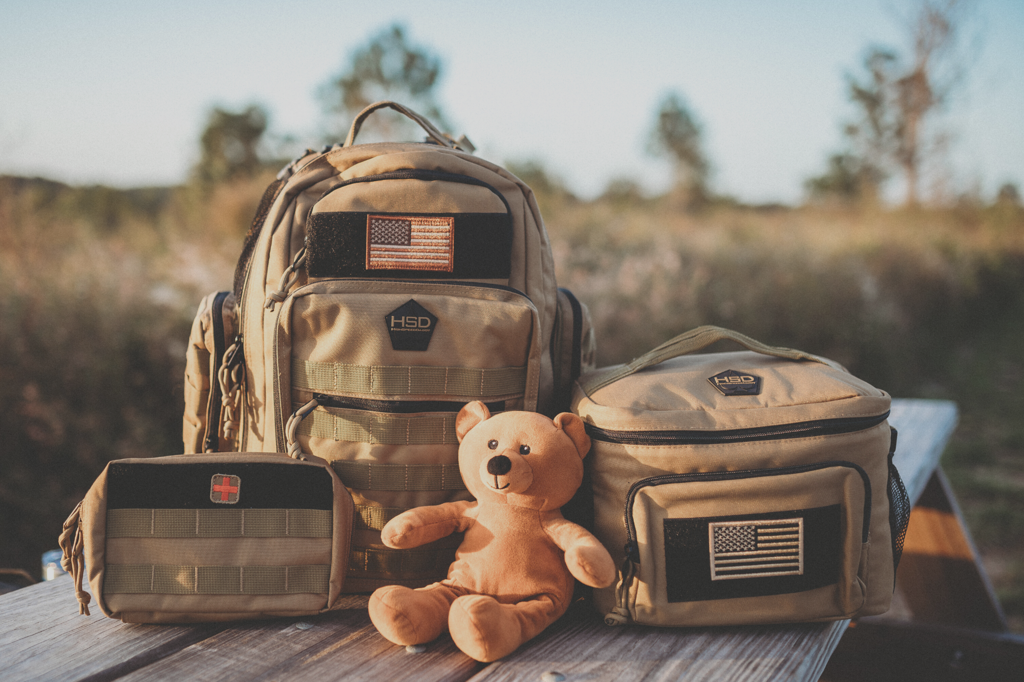 Camouflage backpack and pouches with a teddy bear on a wooden surface outdoors.