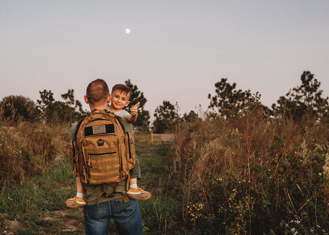Man with a child in a field at dusk, both wearing tactical backpacks.