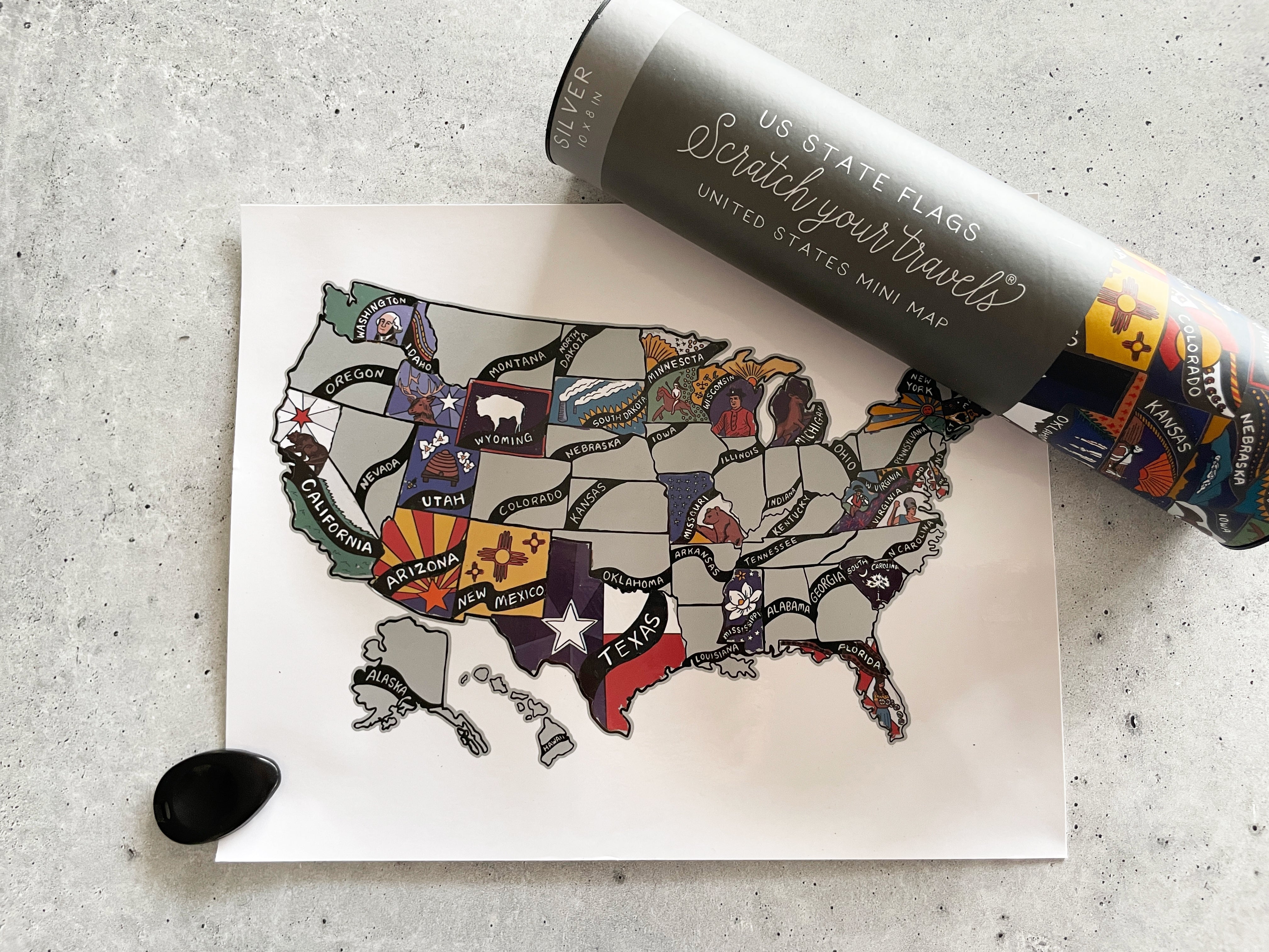 Map of the United States with state flags and a scratch-off travel map on a gray surface.