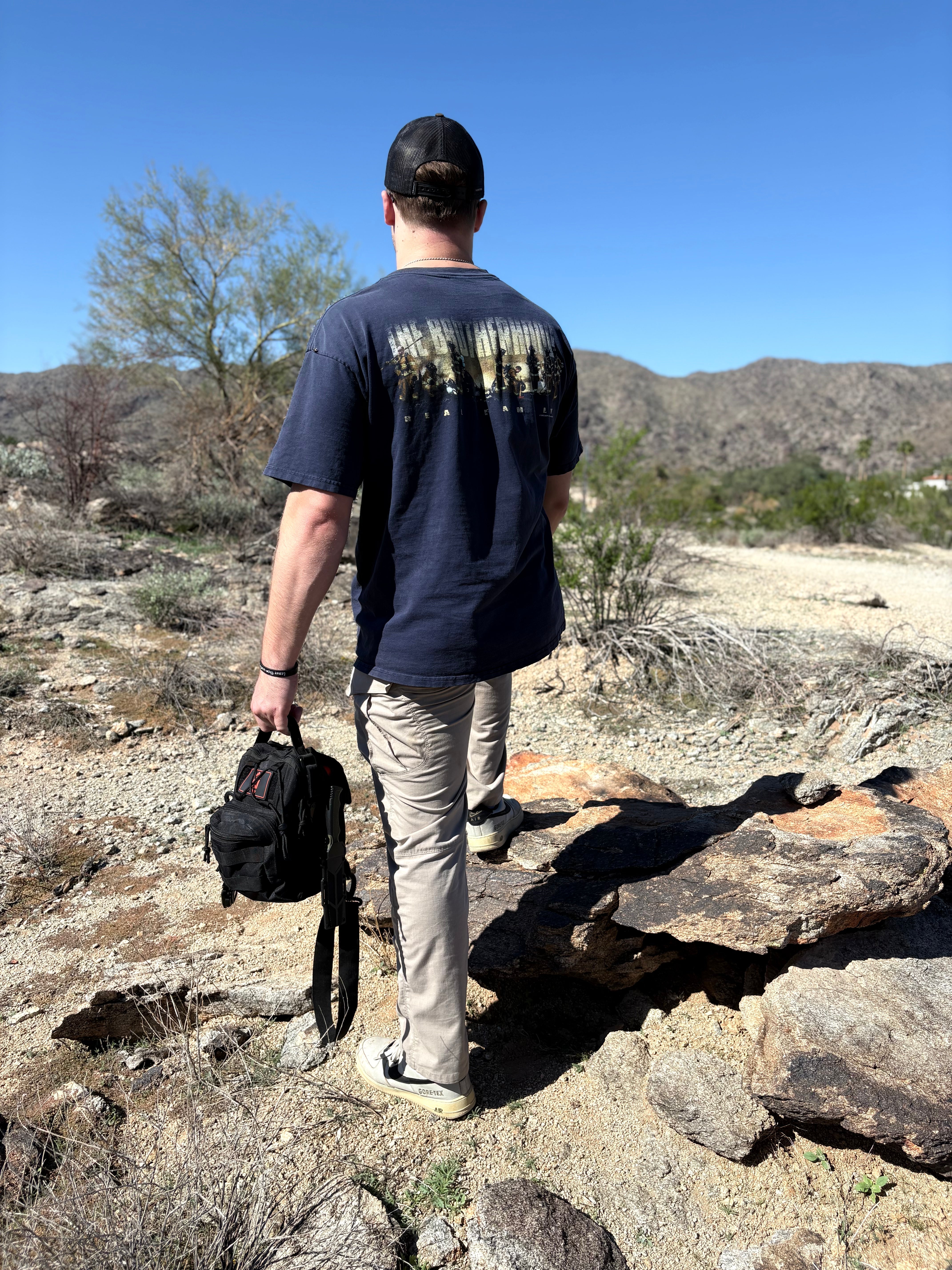 Person standing on a rocky landscape holding a backpack, wearing a dark blue t-shirt with text on the back.