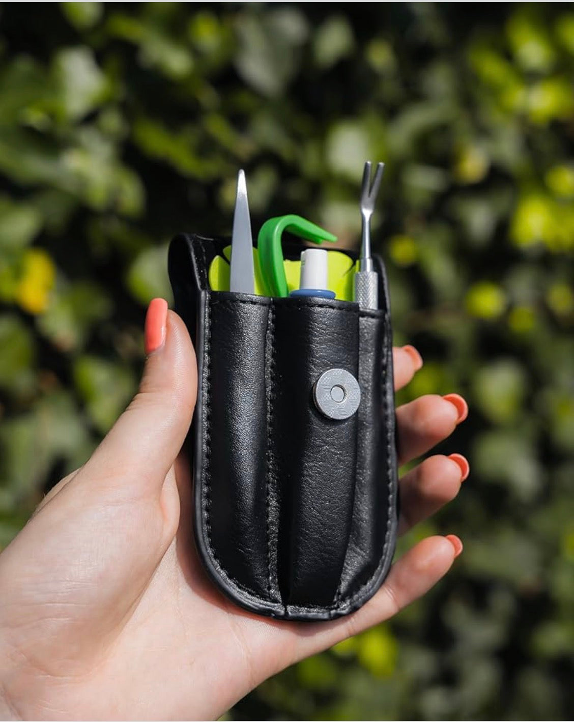 Black leather tool pouch with garden tools held by a hand against a blurred green background