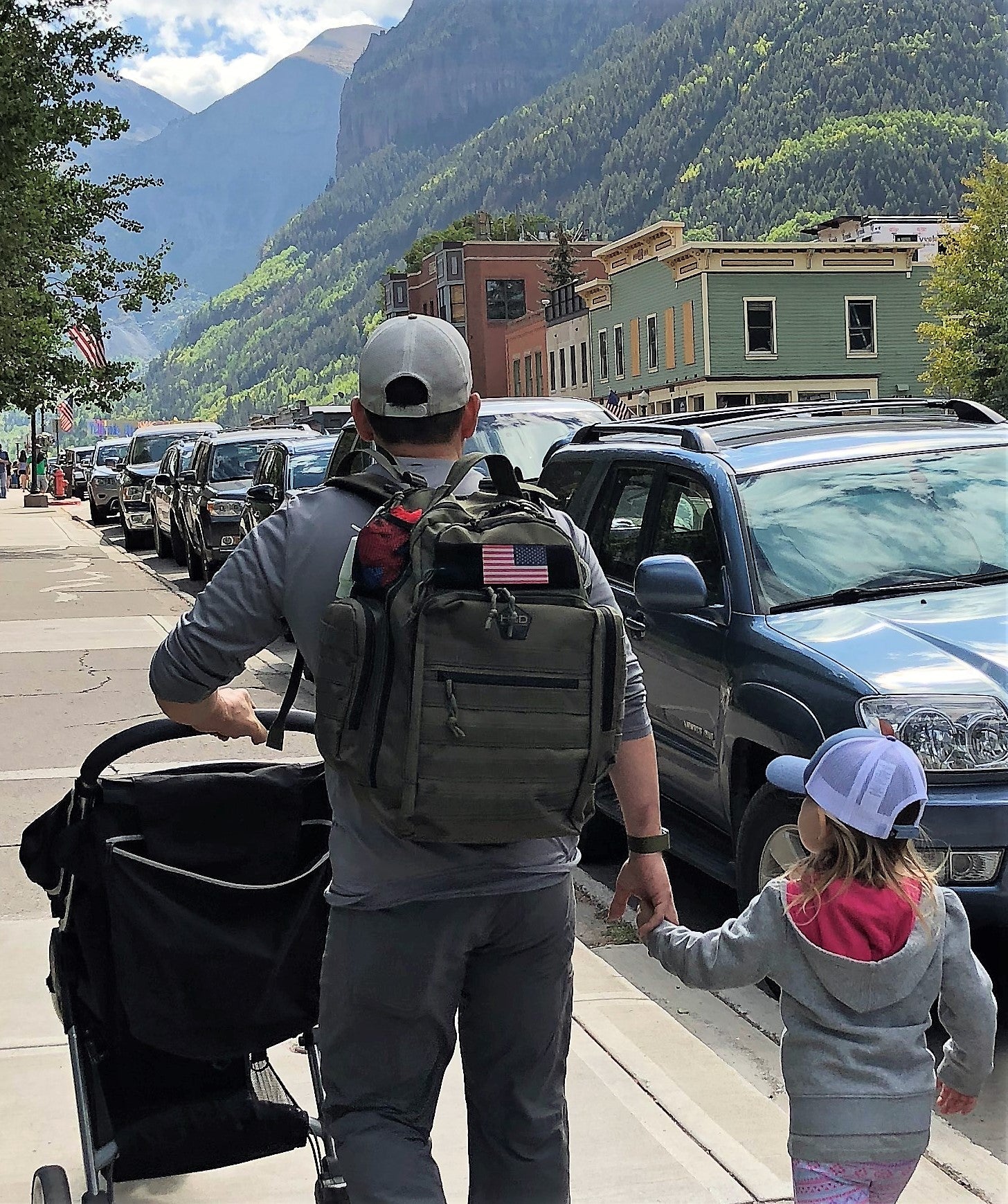 Man with a backpack and child holding hands on a street with mountains in the background