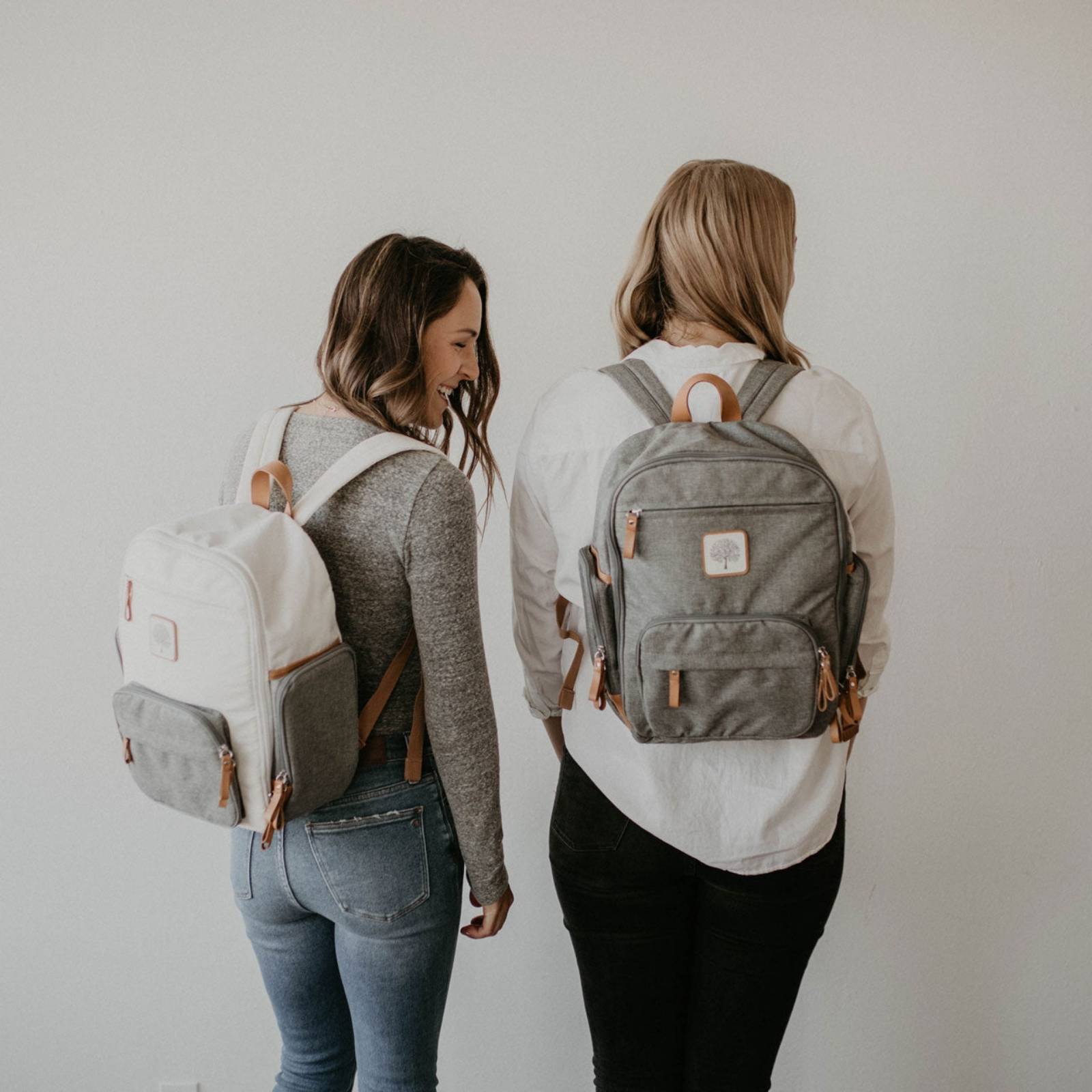 Two women wearing gray backpacks with visible brand logos on a plain background