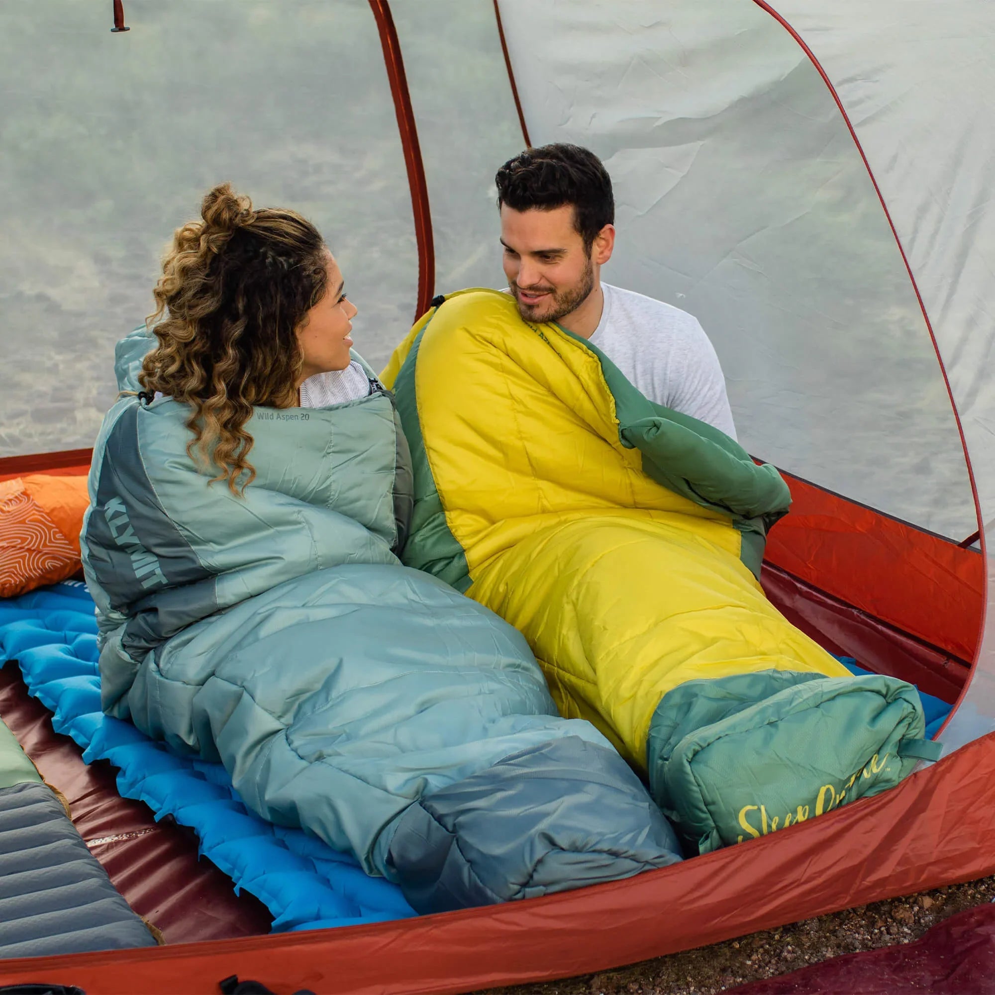 Two people lying in sleeping bags inside a tent, one in green and the other in yellow.