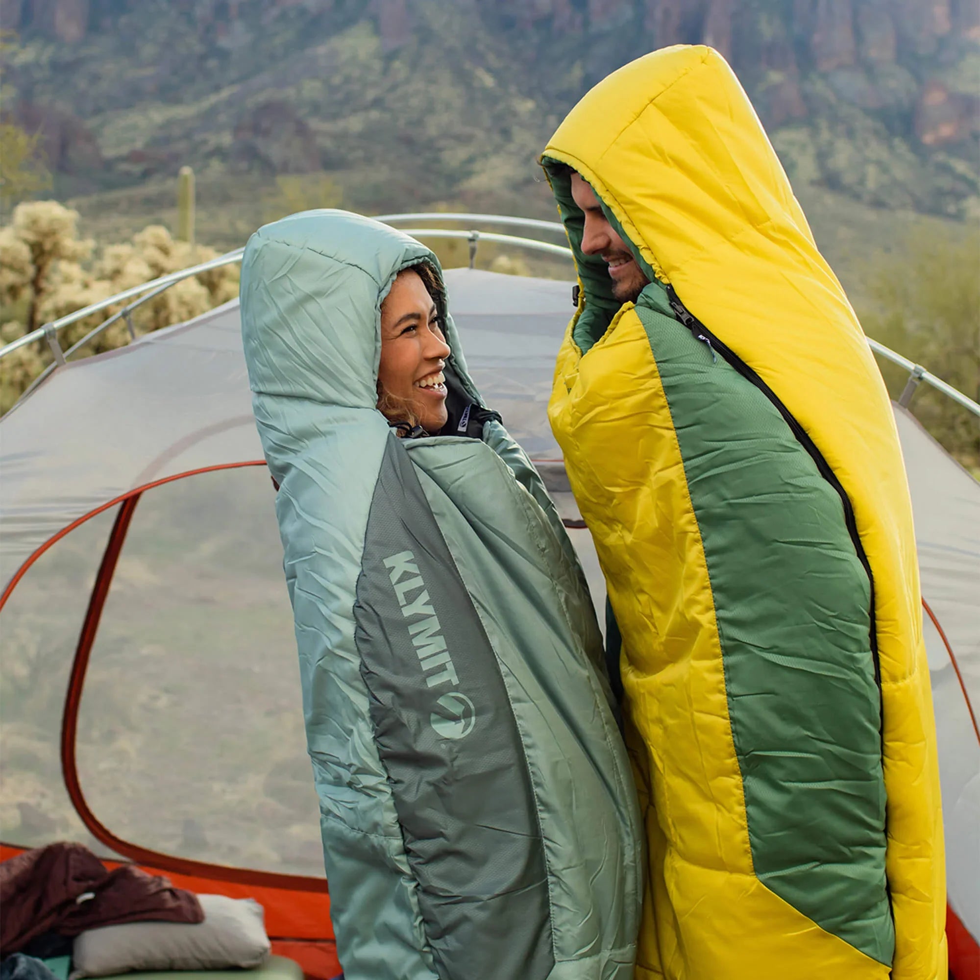 Two people in green and yellow sleeping bags standing near a tent with a mountainous landscape in the background.