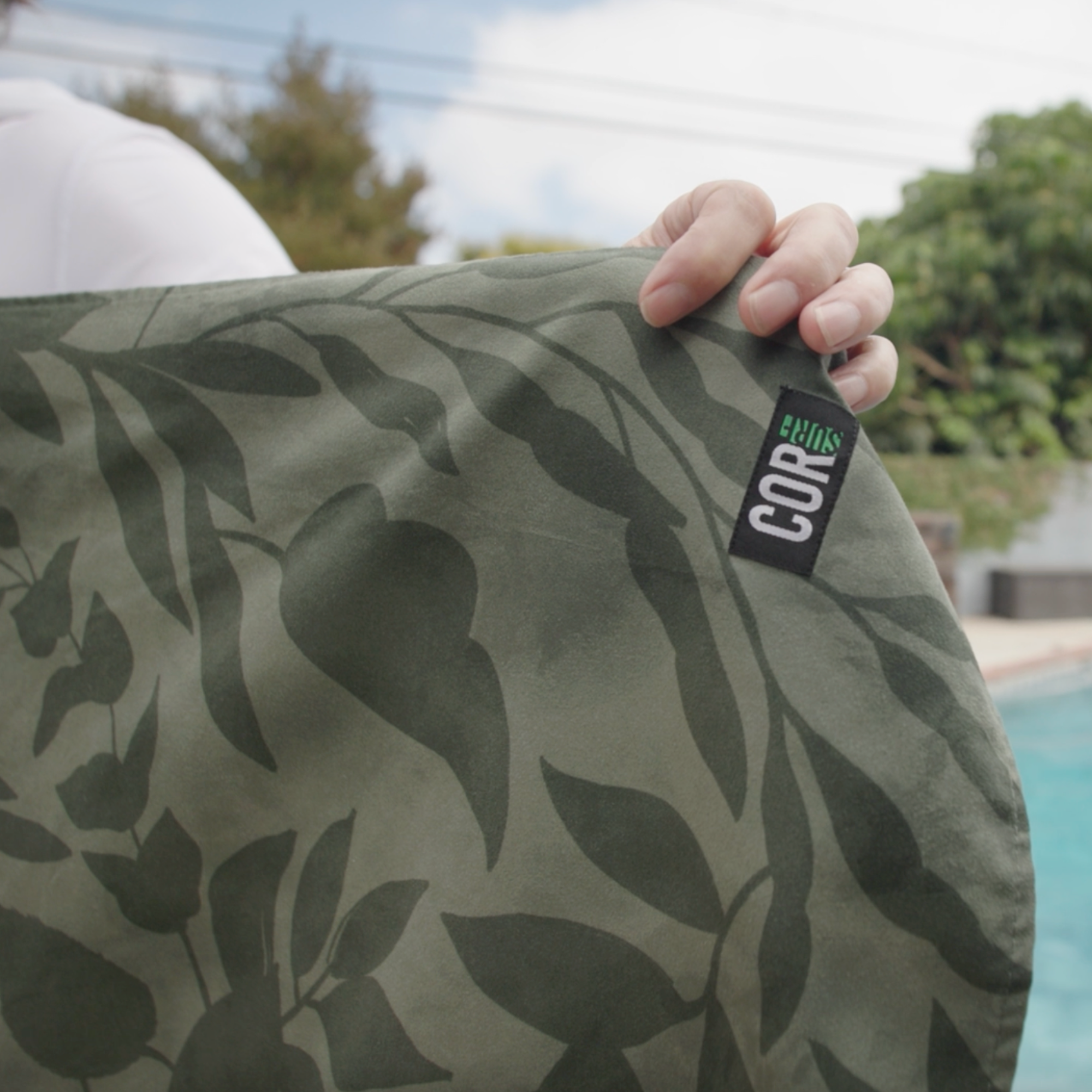 Green leaf-patterned towel with a visible brand logo held by a person outdoors.