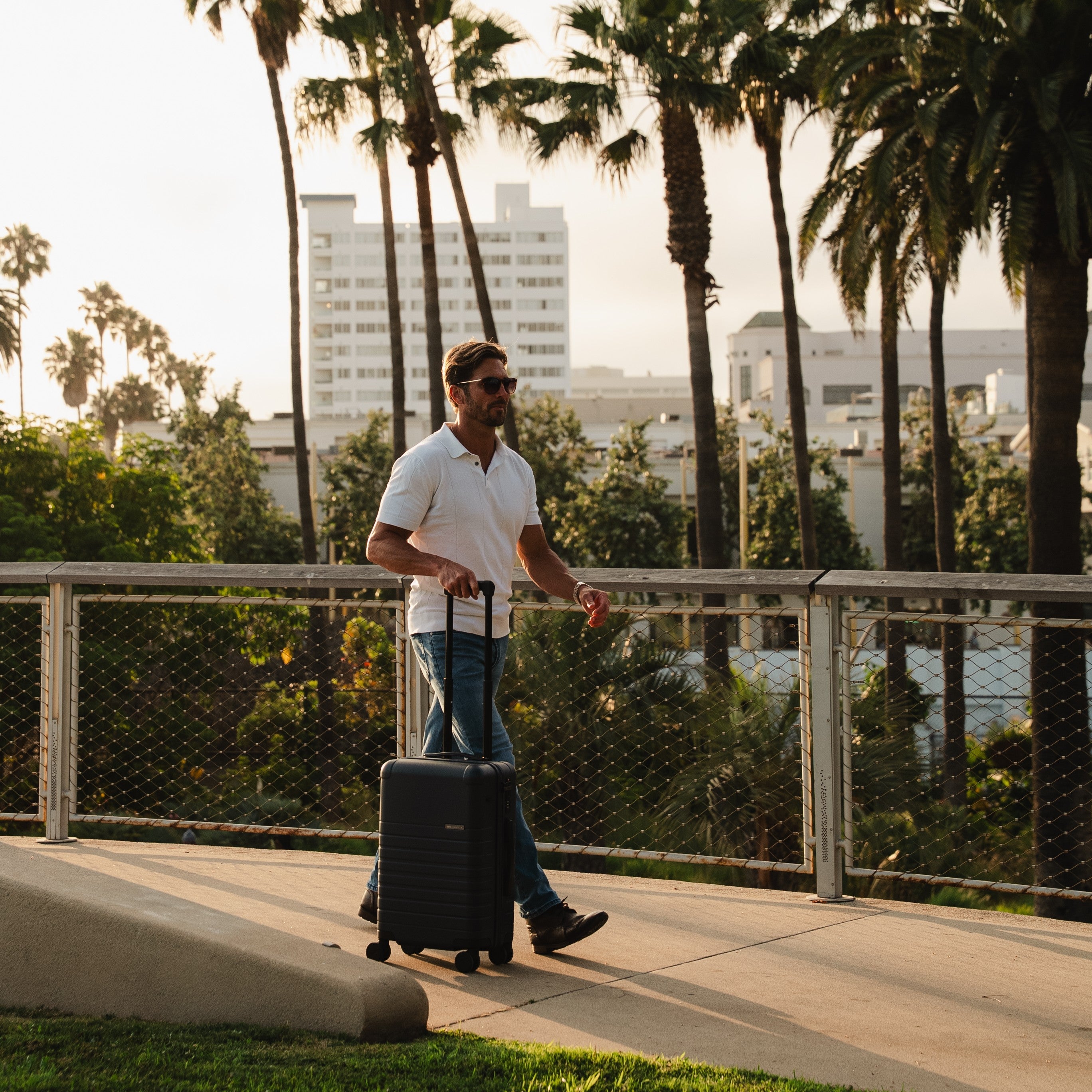 Man with a suitcase walking on a sidewalk with palm trees and buildings in the background