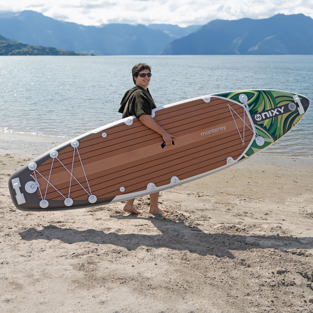 Person holding a large inflatable paddleboard on a beach with mountains in the background