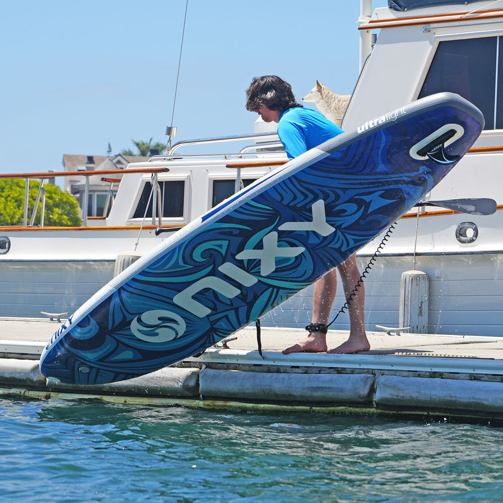Person holding a blue and white wakeboard on a dock with a boat in the background