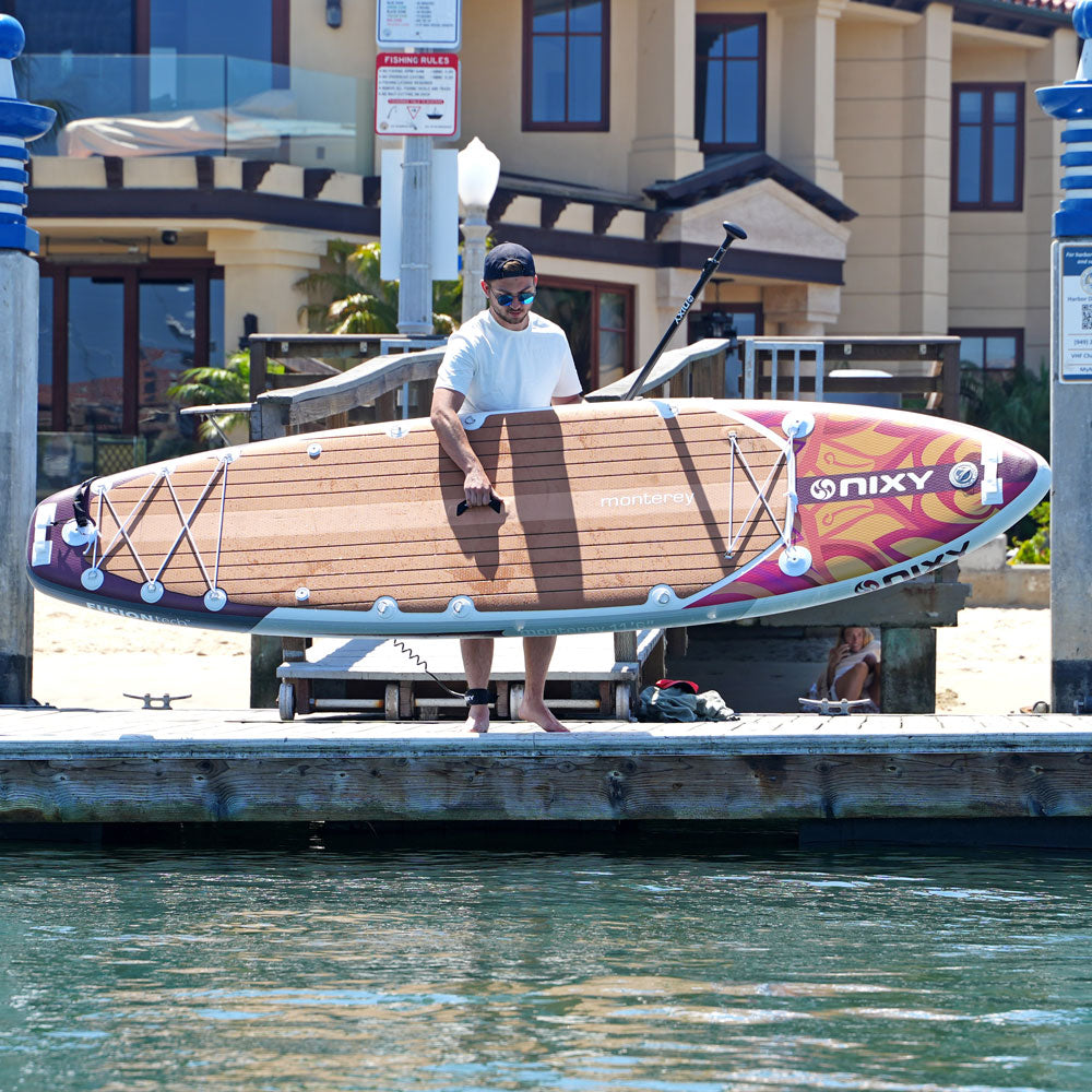 Person holding a large inflatable paddleboard on a dock with a building in the background