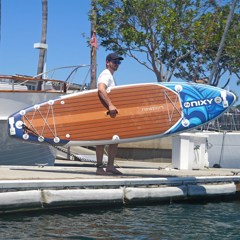 Man holding a wooden and blue Nixy paddleboard on a dock with water and trees in the background.