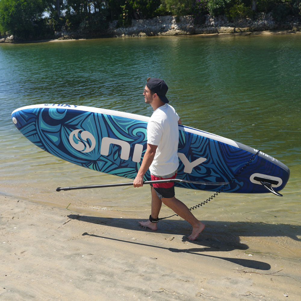 Man carrying a blue and white paddleboard with a logo on a sandy shore.