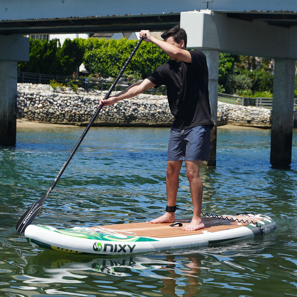 Person paddleboarding on a Nixy board in a body of water with a bridge and greenery in the background.